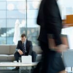 Businessman sitting in modern office lounge using a laptop while colleague walks past, creating dynamic blur. — 3 Benefits of