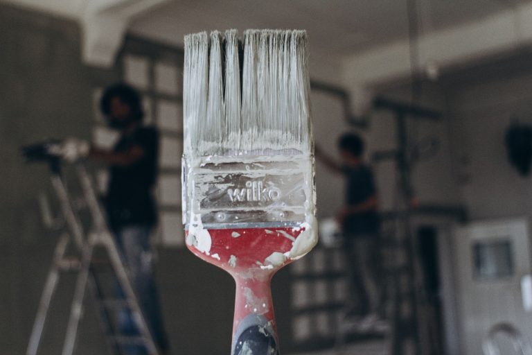 Detailed close-up of a paintbrush with men painting a wall in the blurred background, highlighting renovation work. — Advanta