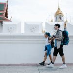 A couple with backpacks walks past stunning Thai temple architecture in an urban setting. — Annual Fly Fishing Trip