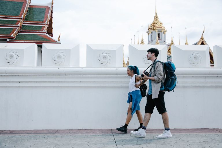 A couple with backpacks walks past stunning Thai temple architecture in an urban setting. — Annual Fly Fishing Trip