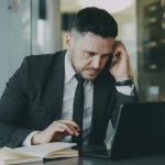 Focused businessman in suit multitasking on phone and laptop in modern office. — Commercial Broker Training 101