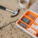 Top view of assorted repair instruments and screws in box near hammer and scotch tape on dirty tile in soft daylight — Differ