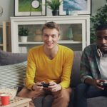 Two friends playing video games together on a sofa, one holding a controller and the other wearing headphones. — Exploring Di