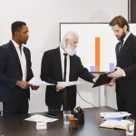 Three businessmen discussing documents in a conference setting with a presentation screen. — Have You thought of Using Entert