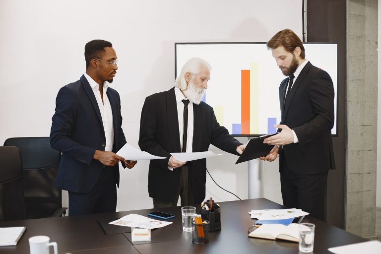 Three businessmen discussing documents in a conference setting with a presentation screen. — Have You thought of Using Entert