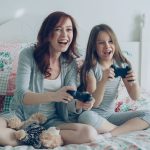 Happy mother and daughter playing video games on bed, enjoying fun family time. — Here Are The Pros And Cons For Undergoing S