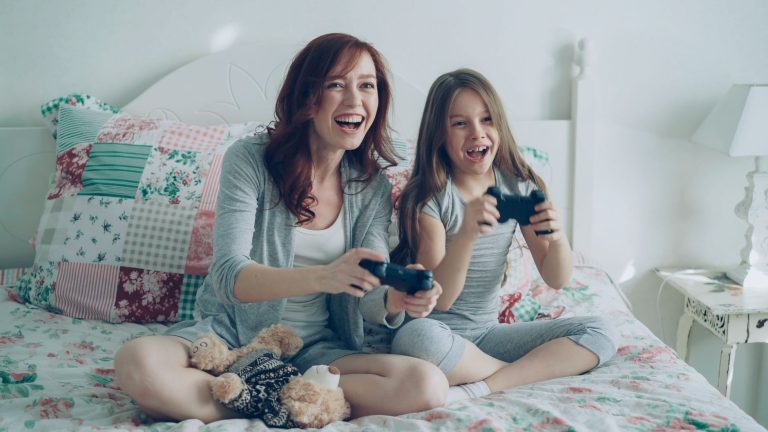 Happy mother and daughter playing video games on bed, enjoying fun family time. — Here Are The Pros And Cons For Undergoing S