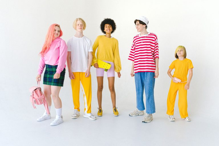 Full body group of positive multiethnic teenager friends with backpack and penny board standing against white wall and lookin