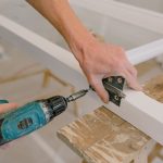 Unrecognizable professional male master using cordless screwdriver to install metal hinge on wooden window in light room duri