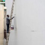 A man on a ladder painting a building's exterior wall with a roller brush outdoors. — Industrial Loft Style Of Room Decoratio