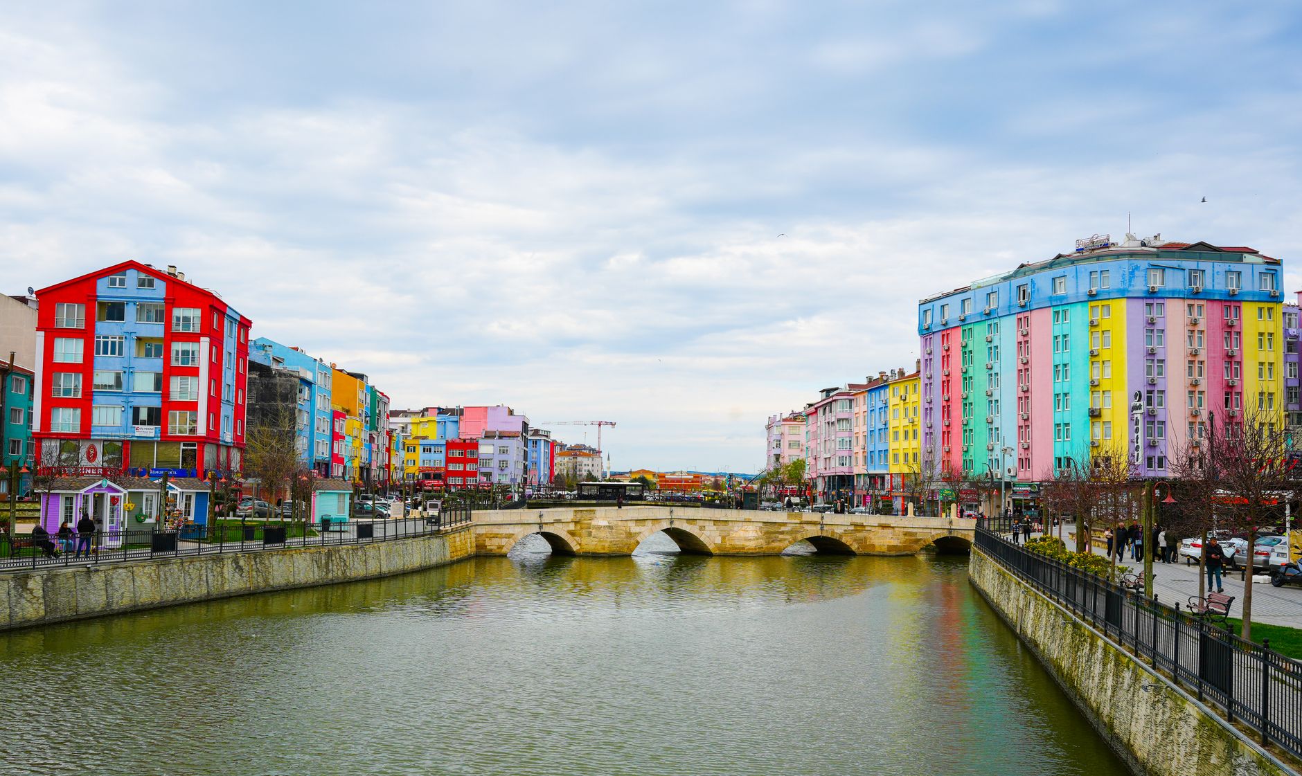 Vibrant cityscape with colorful buildings along a river and stone bridge under a cloudy sky. — Live Action Awaits: Al-Taawoun