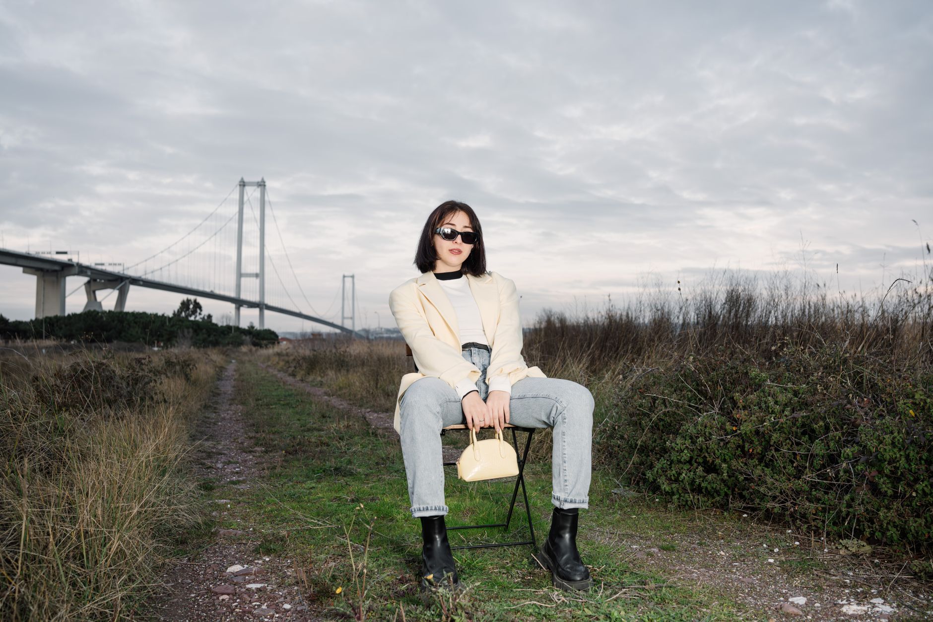 Stylish woman in sunglasses sitting by a bridge, showcasing modern fashion against a natural backdrop. — Raw Cannabis Delight