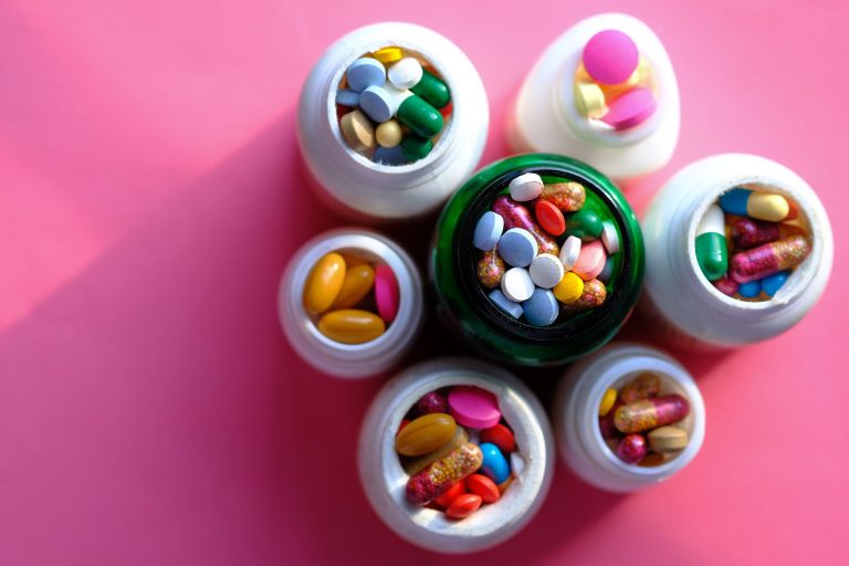 Top view of various medication pills and capsules in containers with bright pink backdrop. — The Cost of Testosterone Therapy