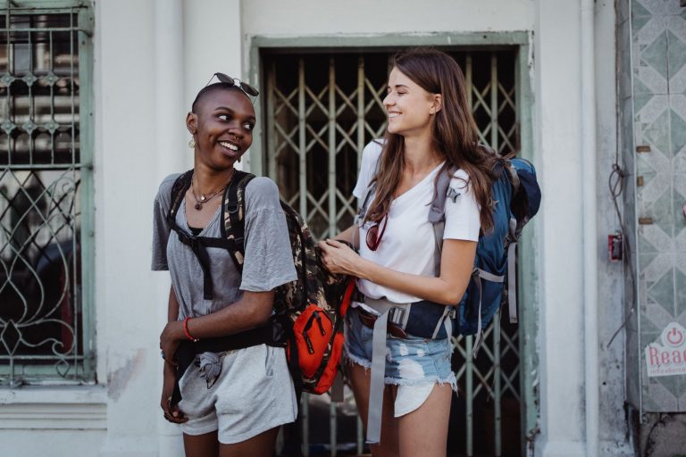 Two young women backpacking and smiling in a lively urban setting, ideal for travel-focused content. — The General Hiring Sol