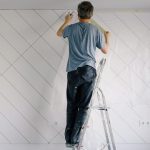 Full body back view of faceless male in shirt and dirty pants repainting wall while standing on ladder in bright empty room i