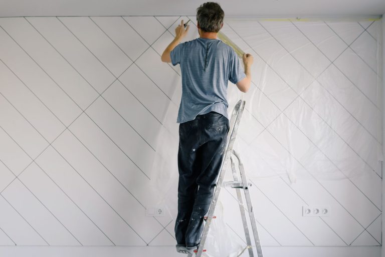 Full body back view of faceless male in shirt and dirty pants repainting wall while standing on ladder in bright empty room i