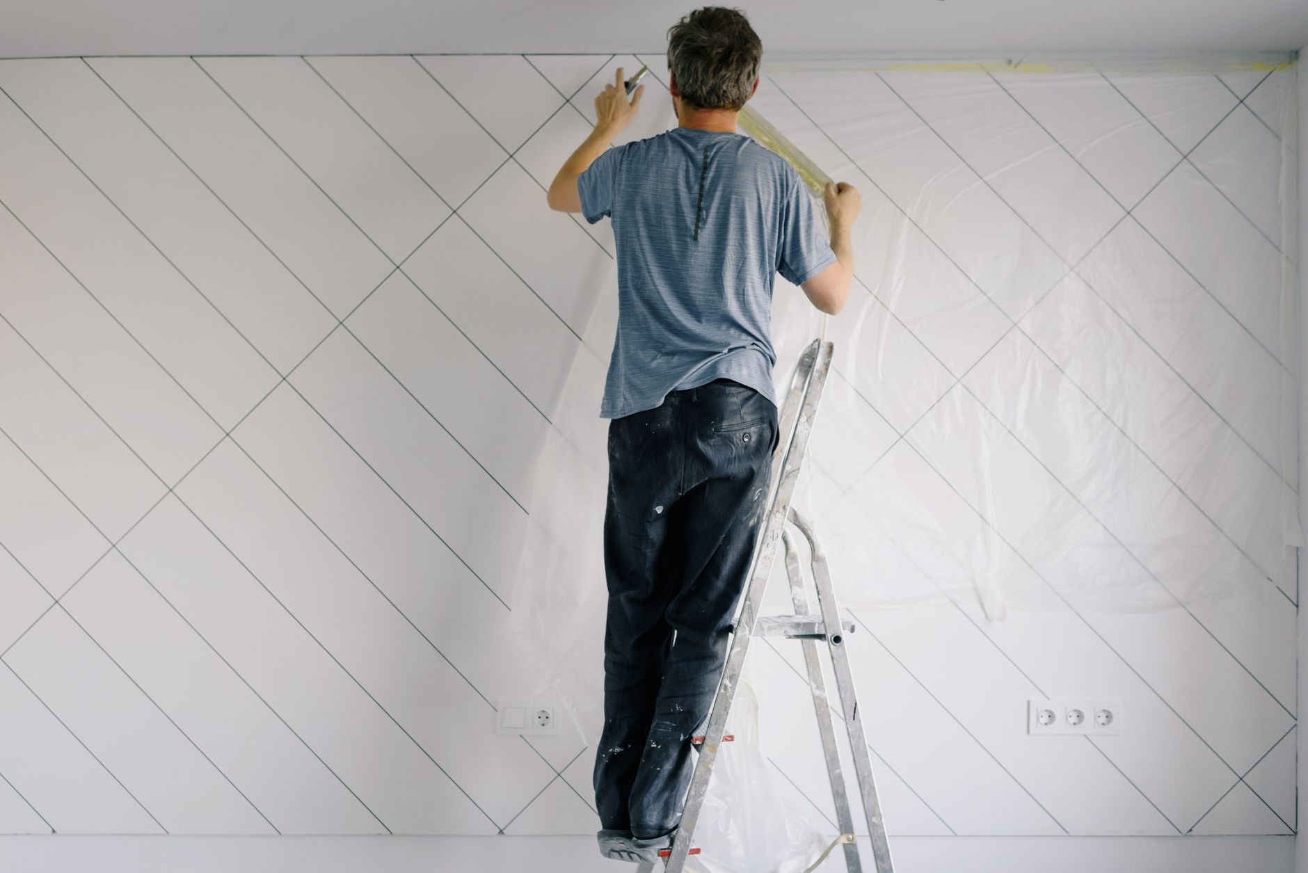 Full body back view of faceless male in shirt and dirty pants repainting wall while standing on ladder in bright empty room i