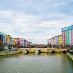 Vibrant cityscape with colorful buildings along a river and stone bridge under a cloudy sky. — U.S. Players Start Instantly a
