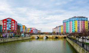 Vibrant cityscape with colorful buildings along a river and stone bridge under a cloudy sky. — U.S. Players Start Instantly a