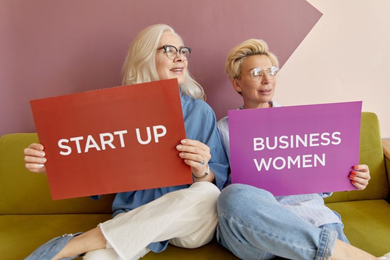 Two women on a sofa holding signs promoting startup and business women. — Understanding the Pros and Cons of Free IPTV