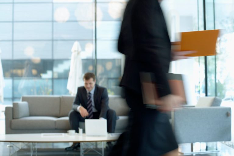 Businessman sitting in modern office lounge using a laptop while colleague walks past, creating dynamic blur. — Unleashing th