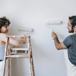 Cheerful adult ethnic couple in aprons looking at each other while painting wall with rollers during renovation process in ho