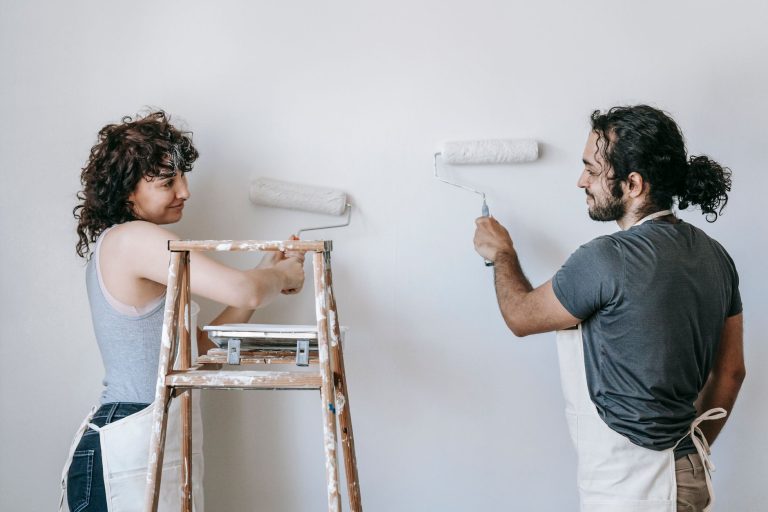Cheerful adult ethnic couple in aprons looking at each other while painting wall with rollers during renovation process in ho