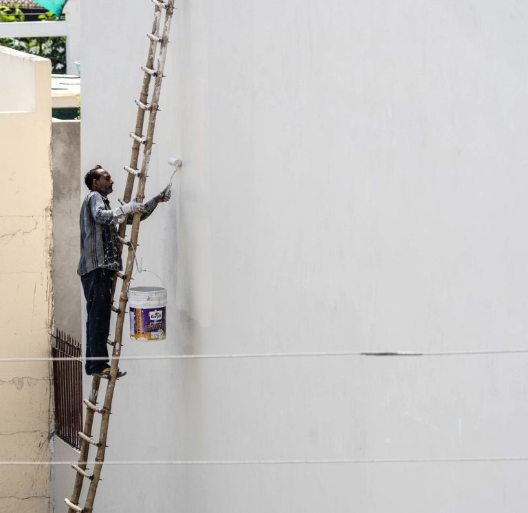 A man on a ladder painting a building's exterior wall with a roller brush outdoors. — Why Choose an eBay Transport Company fo