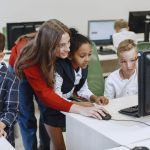 Teacher engaging with diverse children in a technology lesson using computers in a modern classroom. — Why Instruct Your Youn