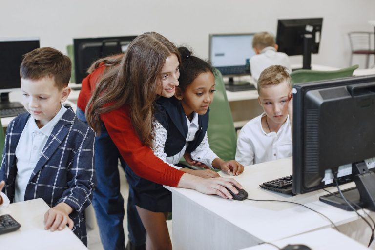 Teacher engaging with diverse children in a technology lesson using computers in a modern classroom. — Why Instruct Your Youn